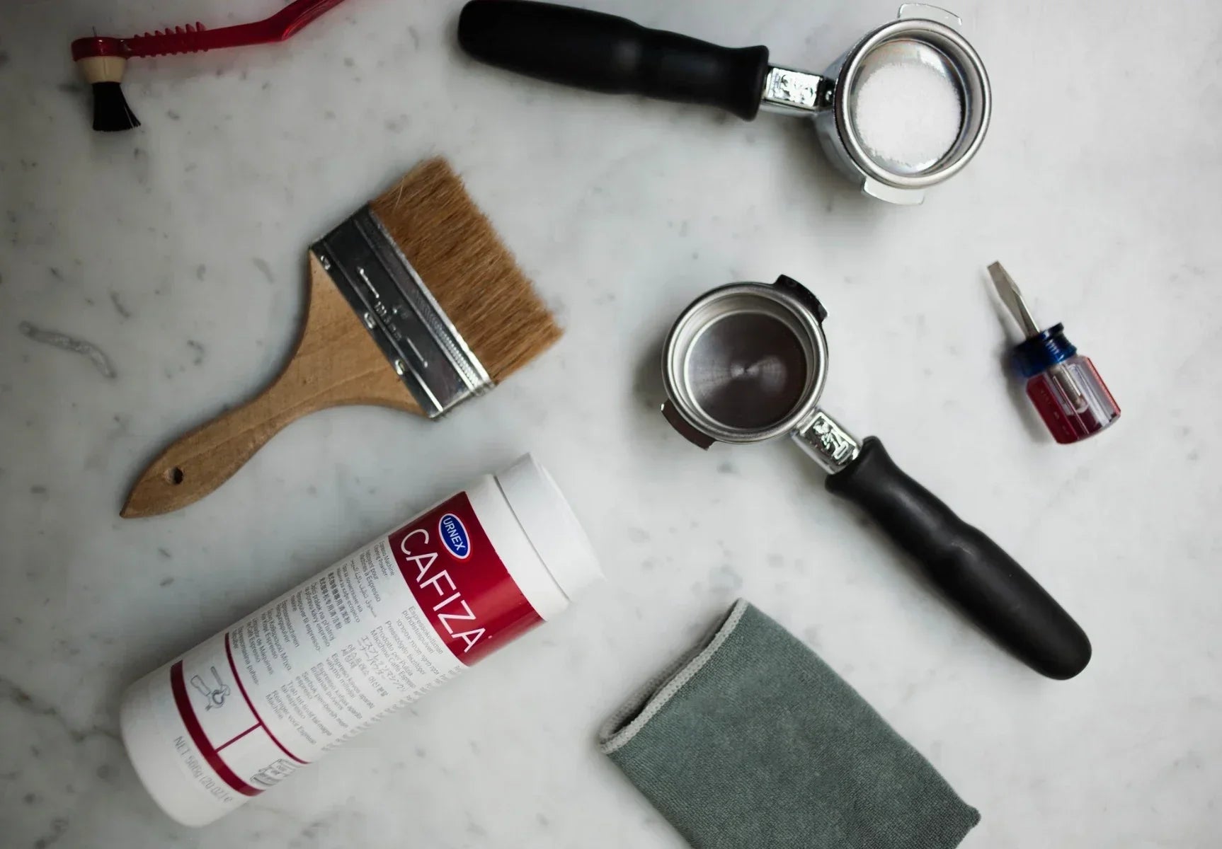 Espresso machine cleaning tools on marble countertop — portafilters, Cafiza cleaner, brush, and cloth used for home coffee machine maintenance.